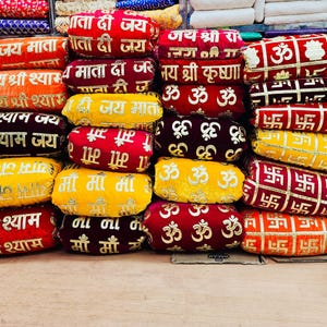 Könnte beinhalten: Ein Stapel bunter, bestickter Kissen. Die Kissen sind in verschiedenen Farben wie Rot, Gelb und Orange gehalten und mit goldenen Texten und Designs versehen. Der Text ist in einer Schriftart gehalten, und die Kissen sind aufeinander gestapelt.