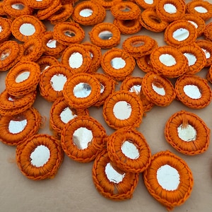 Puede incluir: Una colección de parches circulares bordados en naranja con pequeños espejos plateados reflectantes en el centro. Los parches están densamente dispuestos, mostrando el intrincado trabajo de hilo y las superficies reflectantes.
