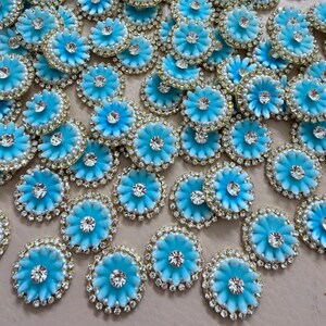 May include: A collection of light blue flower-shaped embellishments. Each embellishment features a central rhinestone, surrounded by a ring of smaller rhinestones and a gold-colored border. The petals are a lighter shade of blue.