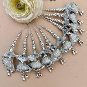 May include: A collection of silver tassels with beaded accents and delicate, fan-shaped embellishments. Each tassel features a cluster of small, dangling charms. A large, cream-colored flower rests in the background, adding a touch of elegance.