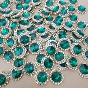 Op de afbeelding: Een verzameling smaragdgroene strass-versieringen. Elke ronde edelsteen is gezet in een goudkleurig metalen frame, omringd door kleine heldere strass-steentjes en een witte parelachtige rand. De juwelen zijn verspreid over het oppervlak.
