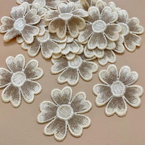 Pode incluir: Um conjunto de patches de flores bordadas. Cada patch apresenta um esquema de cores bege e castanho claro com um design detalhado de pétalas e um centro circular. Os patches estão espalhados sobre uma superfície castanho claro.