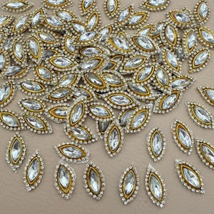 May include: A collection of sparkling, leaf-shaped embellishments. Each piece features a clear, oval-cut rhinestone center, surrounded by a gold-toned metal frame and a border of small, clear rhinestones. These decorative elements are ideal for crafting and embellishment projects.
