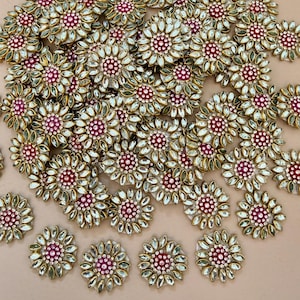 May include: A collection of gold-toned, flower-shaped embellishments. Each piece features a central red-purple jewel surrounded by clear, petal-shaped stones and a gold-colored frame. The embellishments are scattered across a neutral background.
