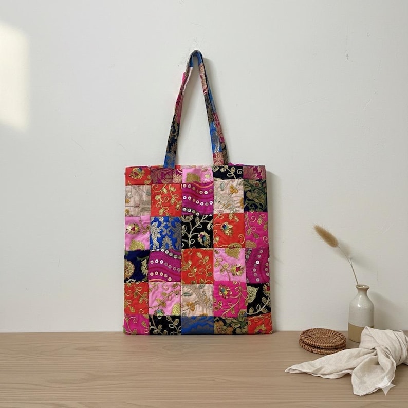 May include: A patchwork tote bag with a colorful design. The bag features a variety of fabrics in squares of pink, red, blue, and gold. The handles are a patterned fabric. The bag is sitting on a wooden surface.