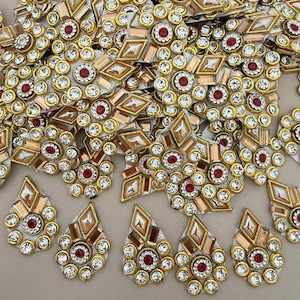 May include: A collection of teardrop-shaped decorative patches featuring gold-toned metallic frames, clear faceted rhinestones, and a central red stone. Each piece is arranged in a symmetrical pattern with geometric accents and circular borders.