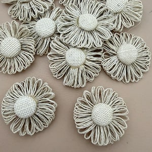 May include: A collection of beige flower-shaped ornaments crafted from natural twine. Each piece features a central circular button covered in textured burlap fabric, surrounded by multiple loops of thin twine forming delicate petals.