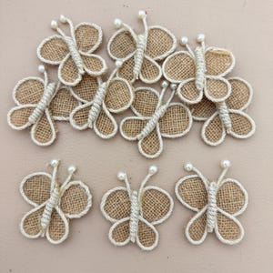 May include: A collection of eight butterfly-shaped ornaments crafted from tan hessian fabric. Each butterfly features a central body and wing outlines made of thick, cream-coloured twisted rope, with two small white beads at the ends of the antennae.