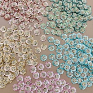 May include: An assortment of decorative floral embellishments in various colors, including pink, gold, teal, and green. Each flower has a central rhinestone and is surrounded by sequins. Ideal for craft projects.