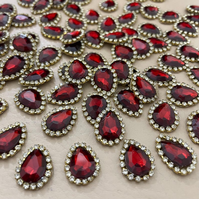 Op de afbeelding: Een verzameling druppelvormige, rode edelsteenversieringen met een goudkleurige rand en een ring van kleine, heldere strass-steentjes. De edelstenen zijn verspreid over een neutrale ondergrond, waardoor hun facetten opvallen.