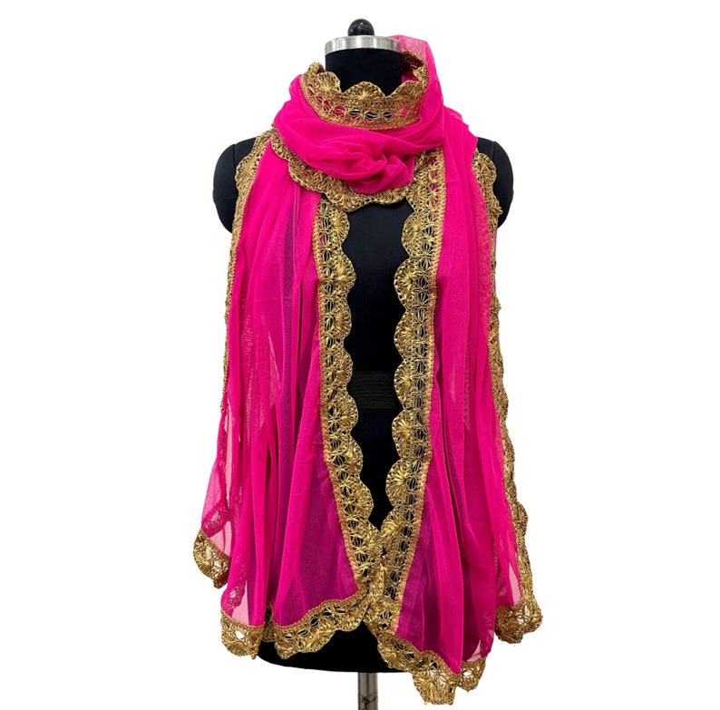 May include: A vibrant pink scarf with intricate gold-colored trim and detailing. The scarf is draped over a black mannequin, showcasing its flowing design and decorative edges. The scarf's fabric appears sheer and lightweight.