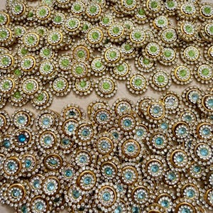 May include: A collection of decorative embellishments with a floral design. Each piece features a gold-colored metal base, a central gemstone, and a ring of clear rhinestones. The gemstones are either light green or light blue.