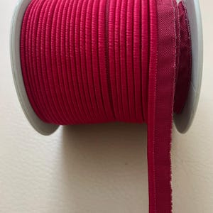 May include: A spool of vibrant fuchsia bias tape. The tape is tightly wound on a gray plastic spool. The edge of the tape is visible, showing a folded and stitched design. The tape is a solid color, with a smooth, slightly shiny texture.
