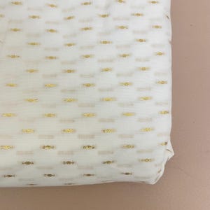 Peut inclure: Gros plan sur un tissu blanc plié avec un motif répété d'accents métalliques dorés. Les accents dorés sont de petits rectangles horizontaux avec un motif texturé. Le tissu semble léger et adapté à l'habillement ou à la décoration intérieure.