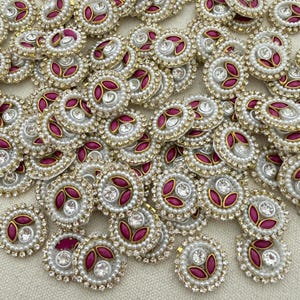Op de afbeelding: Een verzameling ronde geborduurde patches met een centrale heldere strass-steen omringd door drie magenta bloemblaadjes, allemaal omlijst door een rand van kleine witte kunstparels en piepkleine zilveren metalen kraaltjes.