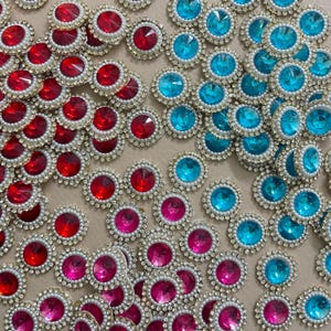 May include: A collection of decorative embellishments in red, pink, and turquoise. Each circular embellishment has a central gemstone, surrounded by small pearls and a border of clear rhinestones. Suitable for craft projects.