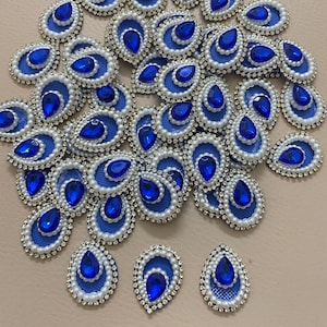 Op de afbeelding: Een verzameling blauwe, druppelvormige versieringen. Elk stuk heeft een grote, levendige blauwe edelsteen omgeven door een witte kralenrand en een blauwe mesh-achterkant. De versieringen zijn verspreid over een neutraal oppervlak.