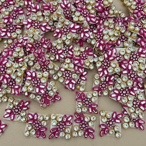 May include: A collection of decorative embellishments. Each square-shaped piece features a central cluster of clear rhinestones surrounded by pink and gold accents. These pieces are ideal for adding sparkle to craft projects.