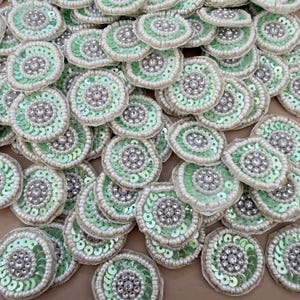 Könnte beinhalten: Eine Sammlung runder Verzierungen. Jedes Stück hat einen zentralen Cluster aus klaren Strasssteinen, umgeben von einem Ring aus silbernen Perlen. Der äußere Rand ist mit hellgrünen Pailletten und einer Bordüre aus weißen Perlen verziert. Ideal für Bastelprojekte.