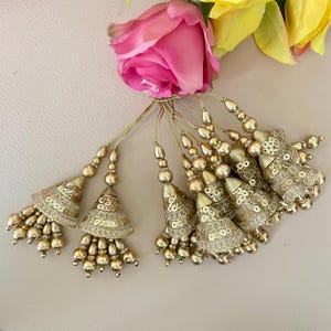 May include: Gold-colored tassels with sequin detailing and beaded accents. The tassels are arranged with a pink rose and yellow flower in the background. The tassels are likely used for decorative purposes.