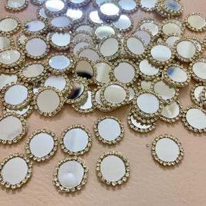 May include: Numerous small, circular mirrored embellishments with a gold-coloured, rhinestone-studded border. The reflective surfaces of the mirrors create a sparkling effect. These decorative elements are scattered across a light pink surface.