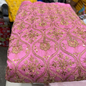 May include: A vibrant pink fabric featuring an intricate gold embroidered pattern of repeating ogee shapes. Each section contains a detailed floral motif accented with small, shimmering gold sequins stitched throughout the design.