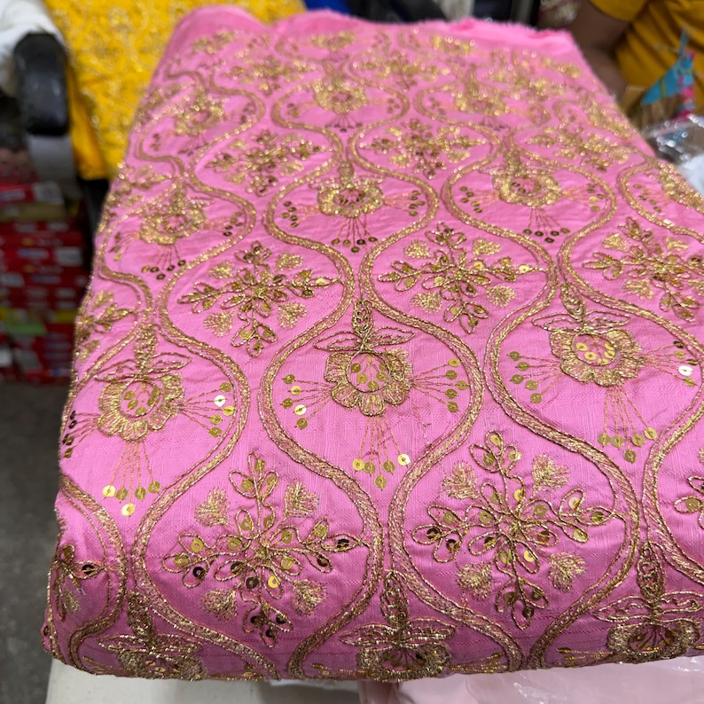 May include: A vibrant pink fabric featuring an intricate gold embroidered pattern of repeating ogee shapes. Each section contains a detailed floral motif accented with small, shimmering gold sequins stitched throughout the design.