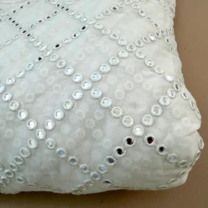May include: Close-up of a white fabric with a repeating pattern of small, round mirrors. The mirrors are arranged in a grid-like design, creating a shimmering effect. The fabric has a soft, textured appearance, suggesting it may be used for clothing or home decor.