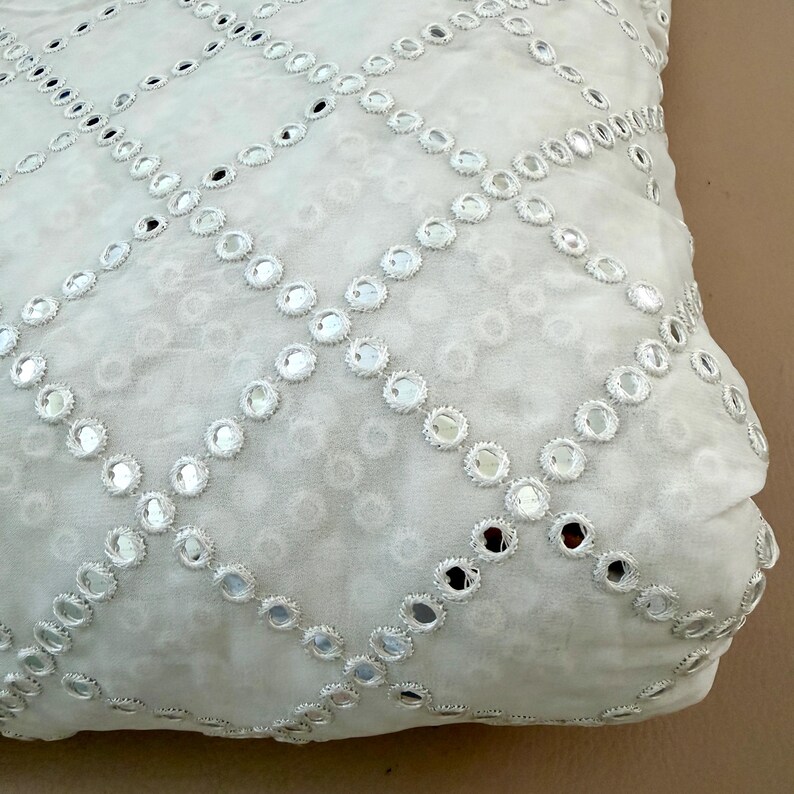 May include: Close-up of a white fabric with a repeating pattern of small, round mirrors. The mirrors are arranged in a grid-like design, creating a shimmering effect. The fabric has a soft, textured appearance, suggesting it may be used for clothing or home decor.