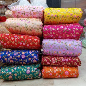 Könnte beinhalten: Ein Stapel bunter Stoffrollen, jede mit floralen Stickereien und Pailletten verziert. Die Stoffe zeigen eine Vielzahl von Farbtönen, darunter Rosa, Gelb, Rot, Blau, Grün, Lila und Orange. Die floralen Muster sind detailliert und lebendig und bilden eine ansprechende Darstellung.