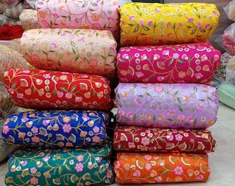 May include: A stack of colorful fabric rolls, each adorned with floral embroidery and sequins. The fabrics display a variety of hues, including pink, yellow, red, blue, green, purple, and orange. The floral patterns are detailed and vibrant, creating a visually appealing display.
