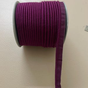 Könnte beinhalten: Eine Spule mit pflaumenfarbener Paspel. Die Zierleiste hat einen tiefvioletten Farbton und scheint aus einem weichen, flexiblen Material zu bestehen. Die Spule ist grau und die Zierleiste ist sauber darum gewickelt. Im Hintergrund ist ein kleines Lineal zu sehen.