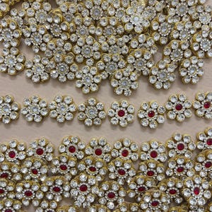 以下が含まれることがあります： 多数の金色の花の装飾品のクローズアップ。各花は、透明で輝くラインストーンと中央の赤い宝石を特徴としています。ジュエリーやアパレルの装飾要素として最適です。