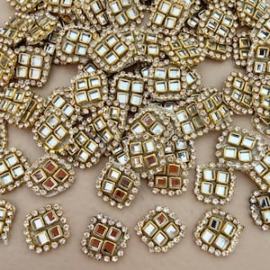 May include: A collection of square-shaped decorative patches featuring four small, reflective mirror-like inserts set within a gold-toned metal frame. Each patch is bordered by a row of tiny, clear, sparkling rhinestones, creating a uniform design.