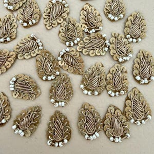 May include: A collection of teardrop-shaped embroidered patches featuring metallic gold thread, intricate coiled wire detailing, and a border of small white beads. Each patch displays a central leaf-like pattern crafted with gold bullion wire.