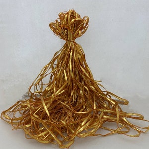 May include: A cluster of gold ribbon, knotted at the top, with numerous strands cascading downwards. The ribbon has a shimmering, metallic finish. The arrangement suggests a decorative element, possibly for gift wrapping or craft purposes.