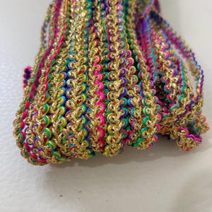 Op de afbeelding: Close-up van kleurrijke decoratieve sierband met een gouden metallic rand. De sierband heeft een herhalend patroon van regenboogkleurige draden in roze, groen, geel en blauw. De gouden rand voegt een vleugje glans en elegantie toe aan het ontwerp.