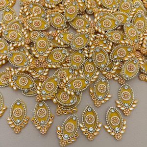 May include: A collection of yellow and gold oval-shaped embellishments. Each piece features a central circular design with a pearl-like border, surrounded by clear, sparkling accents. The embellishments are scattered, showcasing their intricate details.