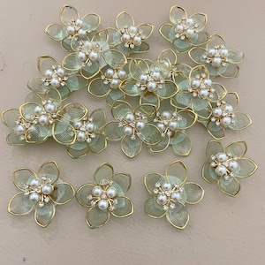 May include: A collection of floral embellishments. Each flower has a pale green translucent petal design with gold-coloured outlines. The centre of each flower features faux pearls and small clear rhinestones. These decorative flowers are ideal for craft projects.