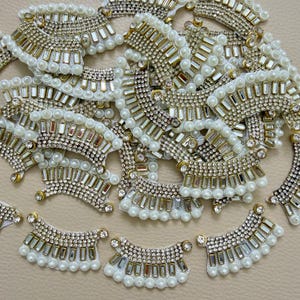 May include: A collection of curved, decorative patches featuring a white base adorned with rows of clear rhinestones, rectangular silver-toned metallic accents, and a border of small white faux pearls arranged in a semi-circular pattern.