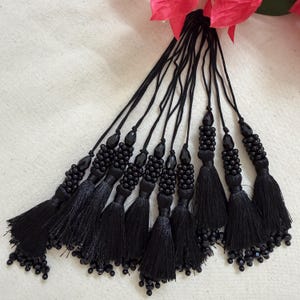 Peut inclure: Un ensemble de pompons noirs suspendus à de fins cordons noirs. Chaque pompon présente un groupe de petites perles noires sur le dessus et une frange dense de fils noirs en bas, avec des perles noires supplémentaires suspendues aux extrémités.