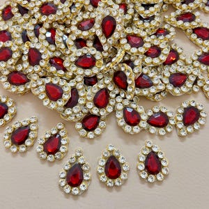 May include: A collection of ornate, teardrop-shaped embellishments. Each piece features a deep red, gem-like center stone surrounded by a halo of clear, sparkling rhinestones. The gems are set in a gold-colored metal base.