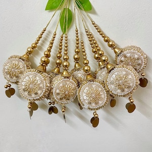 Puede incluir: Un conjunto de borlas decorativas doradas y blancas. Cada borla presenta un diseño circular bordado con lentejuelas y cuentas doradas, acentuado con pequeños dijes dorados en forma de hoja. Las borlas están suspendidas de hilos dorados con cuentas.