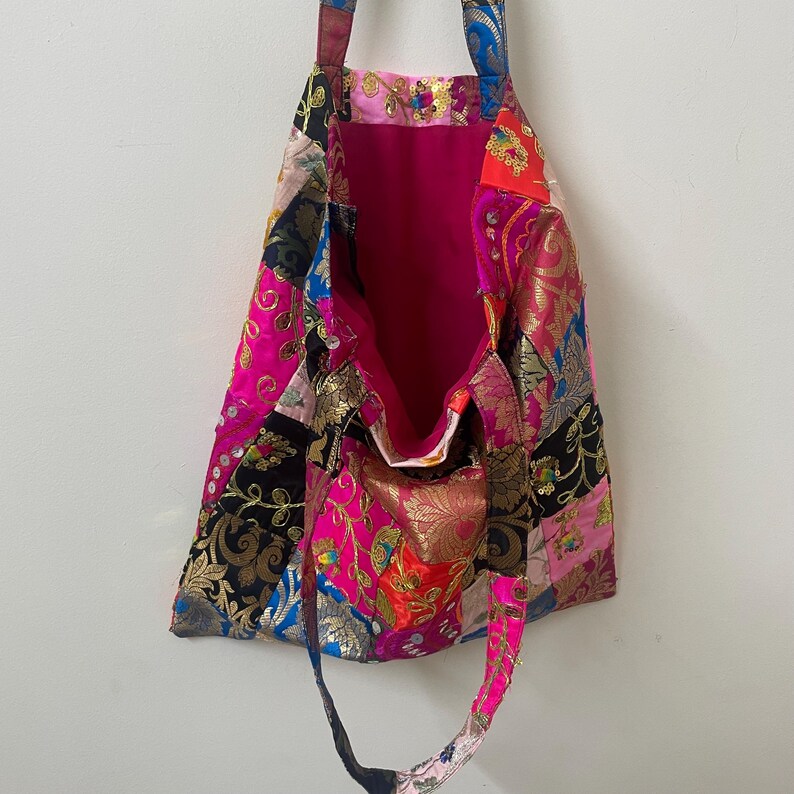 May include: A patchwork tote bag with a vibrant mix of colors and patterns. The bag features a bright pink interior and a variety of fabrics in shades of pink, gold, blue, and black. The bag has two handles and is open at the top.