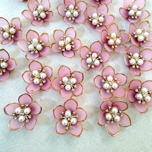 May include: A collection of small, five-petaled pink flower embellishments. Each flower features a gold-toned metal edge and a center cluster composed of three white faux pearls surrounded by tiny, clear, sparkling rhinestones.