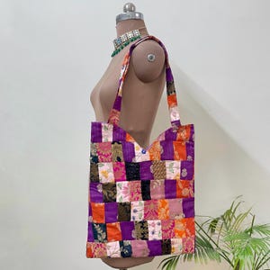 Peut inclure: Un sac fourre-tout patchwork aux couleurs vives, notamment violet, orange et rose. Le sac présente un motif géométrique de carrés de tissu et possède de longues sangles pour être porté à l'épaule. Le sac est présenté sur un mannequin.