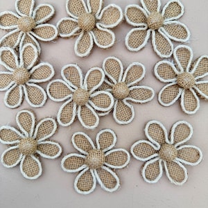 Puede incluir: Un conjunto de nueve adornos con forma de flor hechos de tela de arpillera color canela. Cada flor presenta seis pétalos delineados con un cordón blanco trenzado y un botón central circular de arpillera, creando un aspecto uniforme.