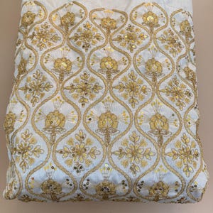 May include: A white fabric featuring an intricate gold embroidered pattern of repeating ogee shapes. Each shape contains a central floral motif accented with small, reflective gold sequins. The embroidery is dense, creating a textured surface.