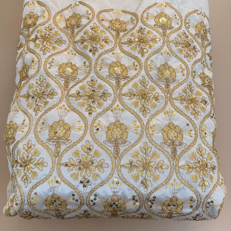 May include: A white fabric featuring an intricate gold embroidered pattern of repeating ogee shapes. Each shape contains a central floral motif accented with small, reflective gold sequins. The embroidery is dense, creating a textured surface.