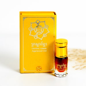 May include: A small bottle of  "GRASSNA KHMER Agarwood Oil" in a gold bottle with a gold cap. The bottle is sitting on a white surface in front of a yellow box with the same text and a silver floral design.